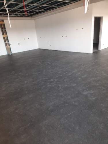 Large open room with dark grey LVT tiles completed, exposed ceiling grid visible during construction