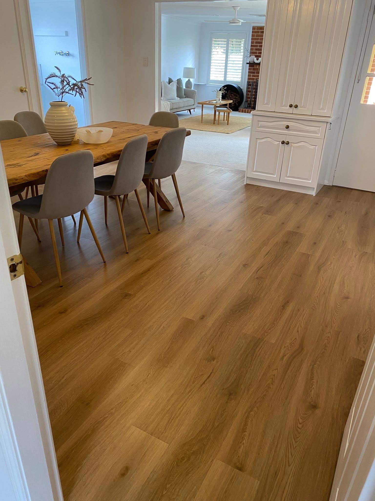 Dining room with warm golden vinyl plank flooring, timber table and grey chairs, looking through to carpeted lounge with exposed brick fireplace